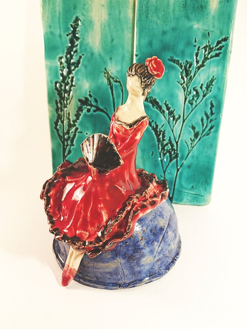May include: A ceramic figurine of a woman in a red dress and a black fan, sitting on a blue and white ceramic base. The figurine is against a green background with a floral design.
