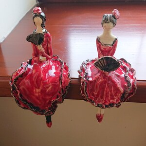 May include: Two ceramic figurines of women in red dresses with black trim and black fans. The figurines are posed in a sitting position with their legs crossed.