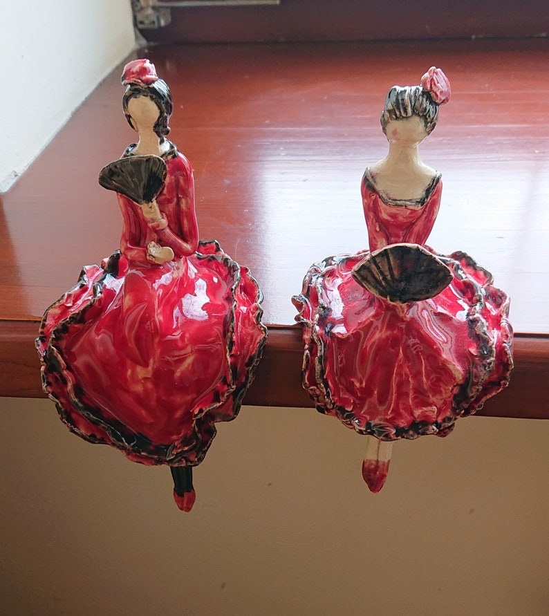 May include: Two ceramic figurines of women in red dresses with black trim and black fans. The figurines are posed in a sitting position with their legs crossed.
