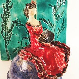 May include: A ceramic figurine of a woman in a red dress holding a fan, sitting on a blue and brown ceramic base.