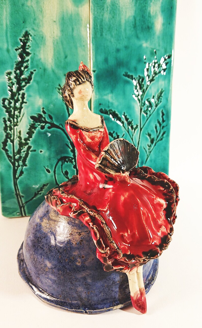 May include: A ceramic figurine of a woman in a red dress holding a fan, sitting on a blue and brown ceramic base.