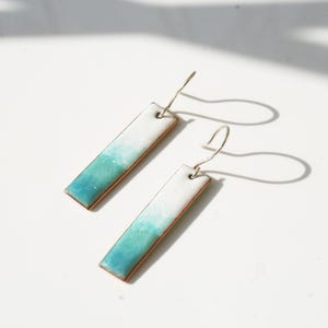 May include: A pair of rectangular dangle earrings with a gradient design. The earrings transition from white to turquoise, with a copper-colored edge and silver-tone ear wires. The earrings are displayed on a white surface.