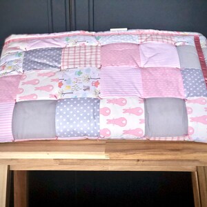 May include: A pink and grey patchwork baby quilt with a pink satin ribbon trim. The quilt is made of various fabrics with different patterns, including bunnies, polka dots, stripes and plaid.