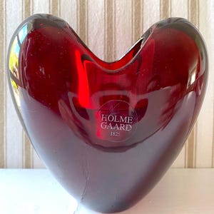May include: A heart-shaped, ruby red glass vase. The vase has a smooth, glossy finish and a wide opening at the top. The text "HOLMEGAARD 1825" is visible on the front. The vase is a decorative item.