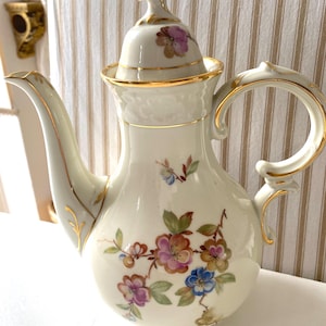 May include: A cream-colored porcelain coffee pot with a lid, handle, and spout, adorned with gold trim. The pot features hand-painted floral designs in shades of pink, blue, and brown. The handle is arched, and the spout is curved.