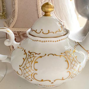 May include: White porcelain teapot with gold filigree detailing and a gold finial on the lid. The teapot has a curved handle and spout, and the design includes ornate patterns. A classic piece for serving tea.
