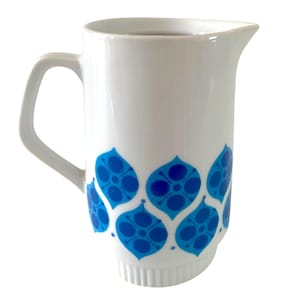 May include: A white ceramic jug featuring a repeating pattern of blue, petal-shaped motifs with dark blue circular centres. The jug has a smooth, cylindrical body, a curved handle on the side, a pouring spout, and a ribbed base detail.