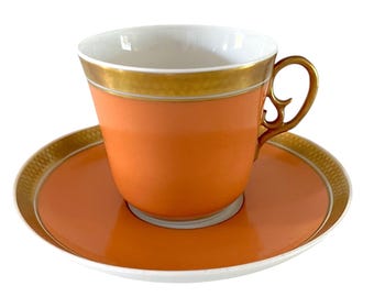May include: A peach-colored teacup and saucer set. The cup has a gold-colored handle and a gold band around the rim. The saucer has a matching peach color with a gold-colored border. The interior of the cup is white.