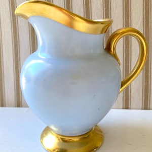 Può includere: Una brocca in ceramica azzurro chiaro con finitura lucida, caratterizzata da bordo, manico e base circolare dipinti in oro. La brocca presenta un corpo arrotondato e un beccuccio curvo, mostrando una superficie liscia e riflettente.