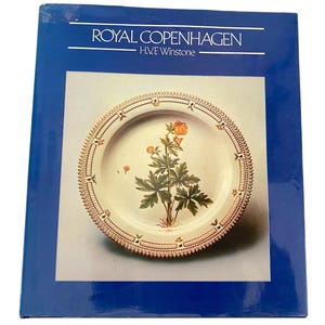 May include: A book cover featuring a Royal Copenhagen plate with a floral design. The plate has a cream-colored background with a detailed illustration of a plant with orange flowers. The plate's rim is decorated with a patterned border.