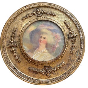 Op de afbeelding: Antiek portret van een vrouw met hoed, geplaatst in een ronde, sierlijke gouden lijst. Het portret heeft zachte kleuren en een vintage esthetiek. De lijst heeft getextureerde details en decoratieve bloemaccenten.