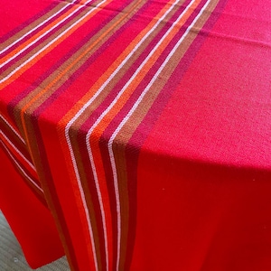 May include: A vibrant red tablecloth featuring a prominent vertical stripe pattern in shades of orange, brown, and white. The fabric has a visible woven texture, and the stripes run along the length of the cloth, creating a bold contrast against the solid red background.