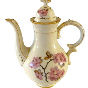 May include: Cream-colored porcelain coffee pot with a floral design. The pot features a curved spout, a rounded body, and a handle. Pink flowers and gold trim adorn the pot, including the lid. A decorative piece for serving coffee or tea.