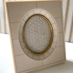 May include: A rectangular off-white picture frame with a central oval opening. The frame features thin black line detailing and a gold-toned metal inner rim. The insert displays a repeating pattern of the letter M in a grid on a light background.