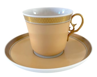 May include: A peach-colored teacup and saucer set with gold trim and a decorative handle. The cup and saucer have a white interior and a gold geometric pattern around the rim. The handle is ornate.