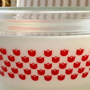 Op de afbeelding: Een witte Pyrex-schaal met een doorzichtig glazen deksel. De schaal is versierd met een patroon van rode tulpen. De schaal heeft twee handvatten en is ontworpen om in te koken en te serveren.