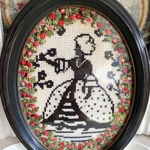 Puede incluir: Una imagen de punto de aguja enmarcada ovalada con una silueta negra de una mujer con un vestido blanco con un borde floral rojo.