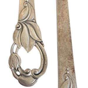 Pode incluir: Um conjunto de talheres de metal prateado composto por um garfo e um cabo de faca. Cada peça apresenta um motivo floral em relevo detalhado na base do cabo, caracterizado por formas de folhas em camadas e linhas curvas com acabamento.