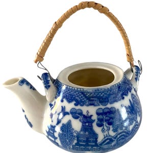 May include: A small, white and blue ceramic teapot with a woven handle. The teapot features a classic blue and white Willow pattern design. The handle is made of woven, light brown material. The spout and rim are white.