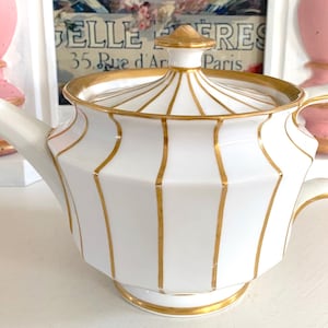 May include: White porcelain teapot with gold trim and a lid. The teapot has vertical gold stripes and a gold rim around the top. The teapot has a handle and a spout.