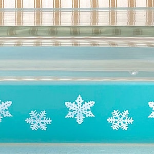 May include: A rectangular turquoise glass baking dish with a clear glass lid. The exterior of the dish features a repeating pattern of white snowflakes printed along the sides. The dish has integrated handles on both ends and a flat base.