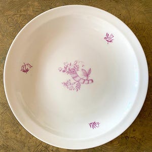 Puede incluir: Plato de cerámica blanco con un borde ancho, decorado con un diseño floral púrpura. El diseño incluye una cornucopia llena de flores y hojas, junto con pequeños acentos florales. El plato es redondo.