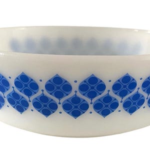 Op de afbeelding: Een witte ovenschaal van melkglas met twee handvatten aan de zijkanten, voorzien van een herhalend blauw geometrisch patroon van gestileerde bladvormen en cirkels rondom de buitenkant. De kom heeft een gladde, ondoorzichtige afwerking.