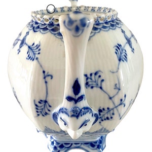 Puede incluir: Una tetera de porcelana blanca con un patrón floral azul y blanco. La tetera tiene un cuerpo redondo con un pico y un asa con forma de cabeza de pájaro. La tetera tiene una tapa con un borde festoneado.
