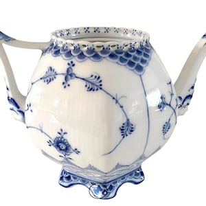 Puede incluir: Una tetera de porcelana blanca con un patrón floral azul y un borde festoneado. La tetera tiene un cuerpo redondo y un pico en el lado derecho. La tetera está sobre un fondo blanco.