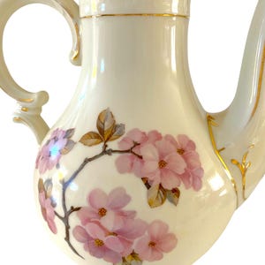 Puede incluir: Cafetera de porcelana color crema con diseño floral. La cafetera tiene un pico curvo, un cuerpo redondeado y un asa. Flores rosas y hojas marrones adornan el cuerpo, con ribetes dorados. Probablemente un objeto vintage o antiguo.