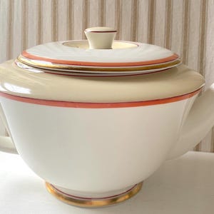 Puede incluir: Una tetera de cerámica blanca con un borde superior color crema, una tapa a juego con un pequeño pomo, y un asa y pico curvos. El diseño está acentuado con finas bandas de color naranja coral y un delicado borde dorado en la tapa y base.