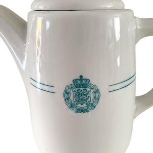 Puede incluir: Una tetera de porcelana blanca con un emblema de color verde azulado en la parte frontal. El emblema muestra un escudo con tres leones coronado por una corona, rodeado por una corona de laurel. Dos finas líneas horizontales rodean el cuerpo.