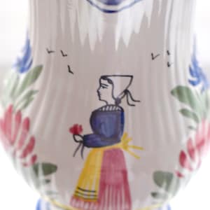 Può includere: Brocca in ceramica bianca con un disegno dipinto raffigurante una donna in abiti tradizionali che tiene un fiore. La brocca ha accenti floreali blu e rossi e un bordo blu. Articoli per la cucina decorativi.