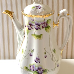 May include: White porcelain coffee pot with a lid, handle, and spout. The pot is decorated with purple violets and green leaves. Gold trim accents the rim, spout, and handle. The pot has a decorative, ornate design.