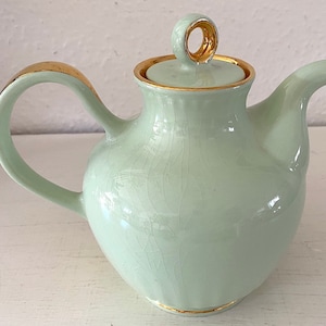以下が含まれることがあります： 金色の取っ手とトリムが付いた、淡い緑色のセラミックティーポット。 ティーポットには、金色のリングハンドルと注ぎ口が付いた蓋が付いています。 ティーポットの表面は、ひび割れのある釉薬効果があります。