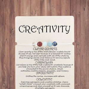 May include: A white card with the word "CREATIVITY" in black lettering. The card features a string of seven crystals: clear quartz, carnelian, aventurine, aquamarine, lapis lazuli, sodalite, and amazonite. The card also includes descriptions of each crystal and its properties.