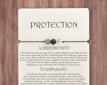 Könnte beinhalten: Eine weiße Karte mit dem Wort "Protection" in schwarzer Schrift oben. Die Karte listet die Eigenschaften von Labradorit, Turmalin und klarem Quarz auf. Die Karte enthält auch ein kleines Armband mit drei Perlen: eine graue Labradoritperle, eine schwarze Turmalinperle und eine klare Quarzperle. Die Karte wurde von CREATIVVO hergestellt.