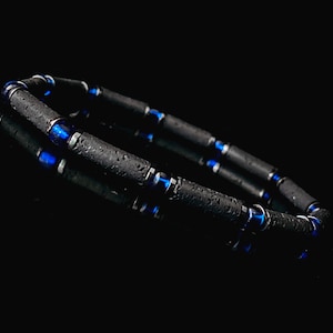 Könnte beinhalten: Ein schwarzes Armband mit blauen Akzenten. Das Armband besteht aus kleinen, runden Perlen, die aufgereiht sind. Die Perlen sind aus einem dunklen, strukturierten Material, möglicherweise Lava-Stein. Die blauen Akzente sind kleine, runde Perlen, die zwischen den schwarzen Perlen verteilt sind.