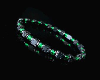 May include: A beaded bracelet featuring a combination of dark gray, square-shaped beads and vibrant green, round beads. The bracelet is displayed against a black background, highlighting its colors and design.