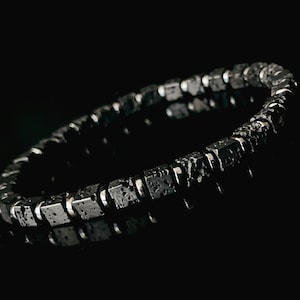 以下が含まれることがあります： 銀色のアクセントが付いた黒い溶岩岩のブレスレット。ブレスレットは正方形のビーズで作られており、手首に着用するように設計されています。
