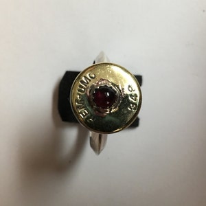 Puede incluir: Un anillo de oro hecho de una vaina de escopeta con una piedra preciosa roja engastada en el centro. La vaina está marcada con "20 GA.  UMC  348".