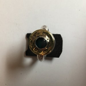 May include: A silver ring with a gold-colored bullet casing set in the center. The bullet casing has a dark blue stone in the center and is engraved with text.
