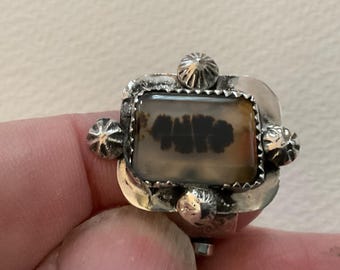 May include: A silver ring featuring a rectangular dendritic agate stone with dark brown fern-like inclusions. The stone is set in a serrated bezel, surrounded by a polished silver frame accented with four small, rounded, ribbed silver beads.