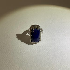 May include: A silver ring featuring a rectangular, deep blue lapis lazuli stone set in a decorative metal frame. The band has a textured, hammered finish, and the stone is secured by a detailed, scalloped metal border surrounding the gemstone.