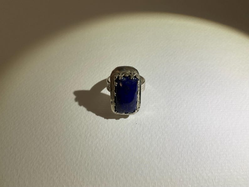 May include: A silver ring featuring a rectangular, deep blue lapis lazuli stone set in a decorative metal frame. The band has a textured, hammered finish, and the stone is secured by a detailed, scalloped metal border surrounding the gemstone.