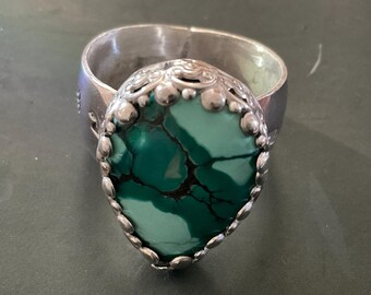 May include: A silver ring featuring a large, pear-shaped turquoise stone with dark matrix veining. The stone is set in a scalloped silver bezel with small decorative metal spheres along the edge, mounted on a wide, polished silver band.