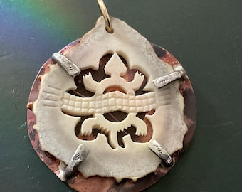 May include: A circular pendant featuring a cream-colored carved bone or antler centerpiece with a lizard motif, secured by four silver-toned metal prongs onto a round, textured copper-colored backing plate with a gold-toned metal jump ring at the top.