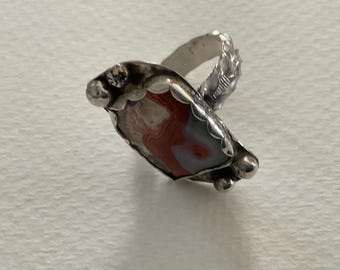 May include: A silver ring featuring a polished, oval-shaped stone with banded layers of red, white, and gray. The metal setting has a scalloped edge, decorative silver spheres at the ends, and a textured, leaf-like pattern along the band.