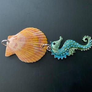 May include: A decorative pendant featuring a natural orange scallop shell attached to a metal seahorse charm. The seahorse is finished in a metallic green tone and is embellished with small, bright blue rhinestones along its curved body.