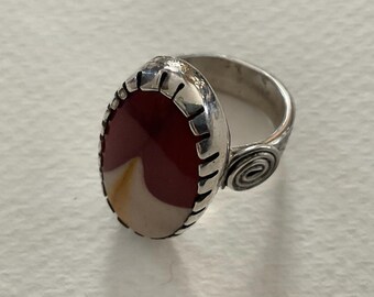 May include: A silver ring featuring an oval-shaped stone with a smooth, multi-colored surface of deep red, cream, and yellow. The metal setting has a notched edge around the stone and a decorative spiral detail on the side of the band.
